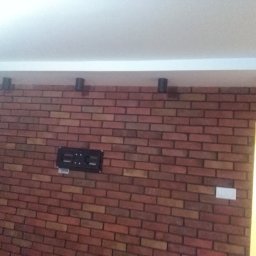 Krispol - Ściana z cegły klinkierowej z zainstalowanymi czarnymi kinkietami sufitowymi i uchwytem na telewizor; widoczny fragment białego sufitu i drzwi.