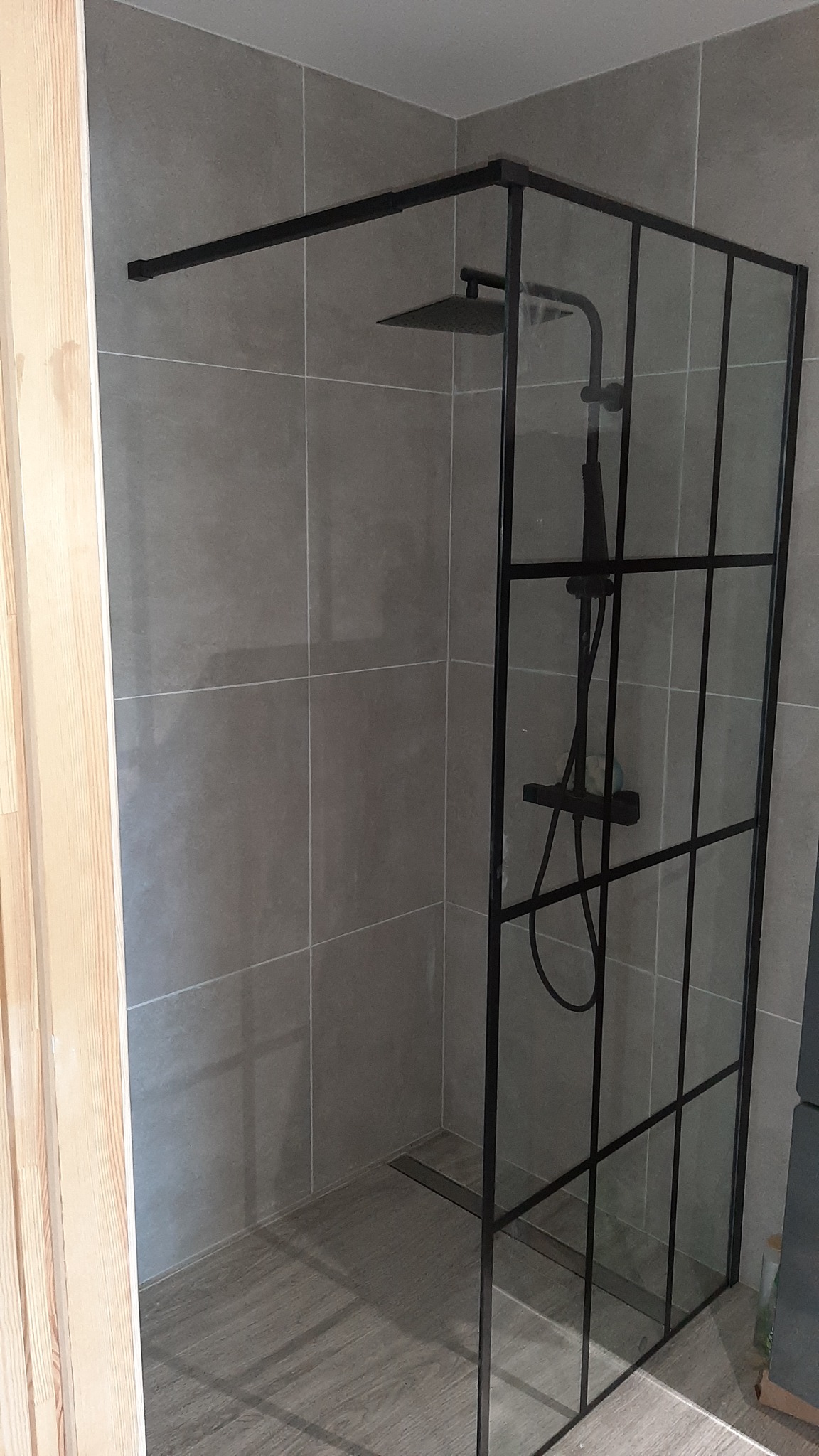Nowoczesna kabina prysznicowa typu walk-in z czarnymi profilami na tle szarych płytek i podłogi imitującej drewno.