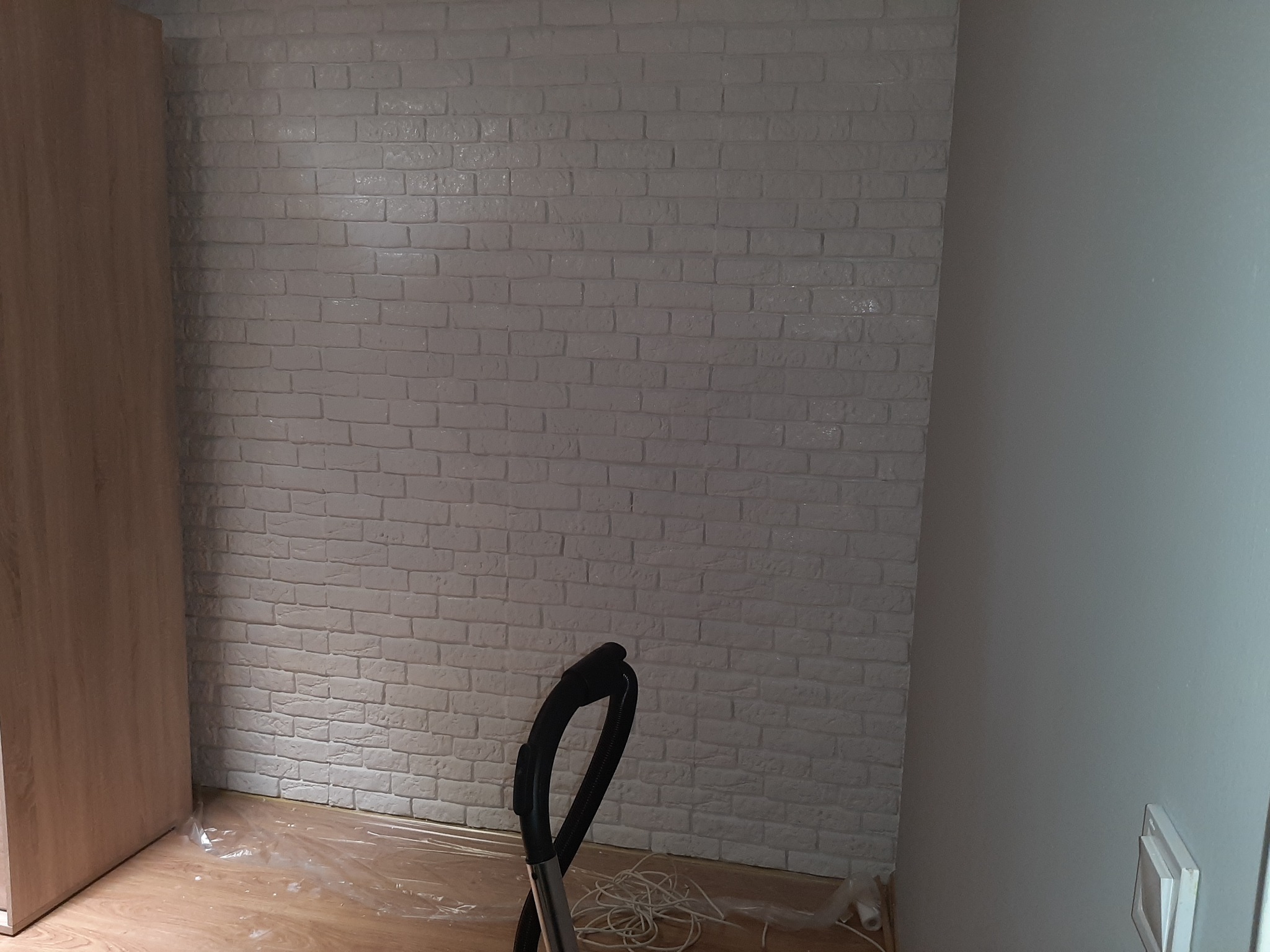Ściana wyłożona białą cegłą dekoracyjną, częściowo zakryta folią ochronną na podłodze, widoczny fragment odkurzacza i szafy, przygotowania do sprzątania po remoncie.