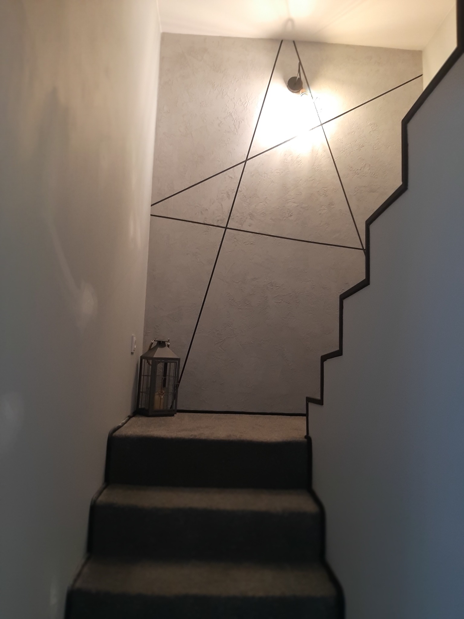 Schody z szarą wykładziną w Ostrołęce, ściana z dekoracyjną strukturą i geometrycznym wzorem z czarnych linii, lampion dekoracyjny u dołu schodów.