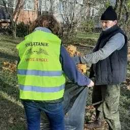 Dwie osoby zbierają opadłe liście do czarnego worka na tle bloku mieszkalnego. Jedna osoba ma na sobie żółtą kamizelkę z napisem 'Sprzątanie' i logo firmy 'Wite Angel'.