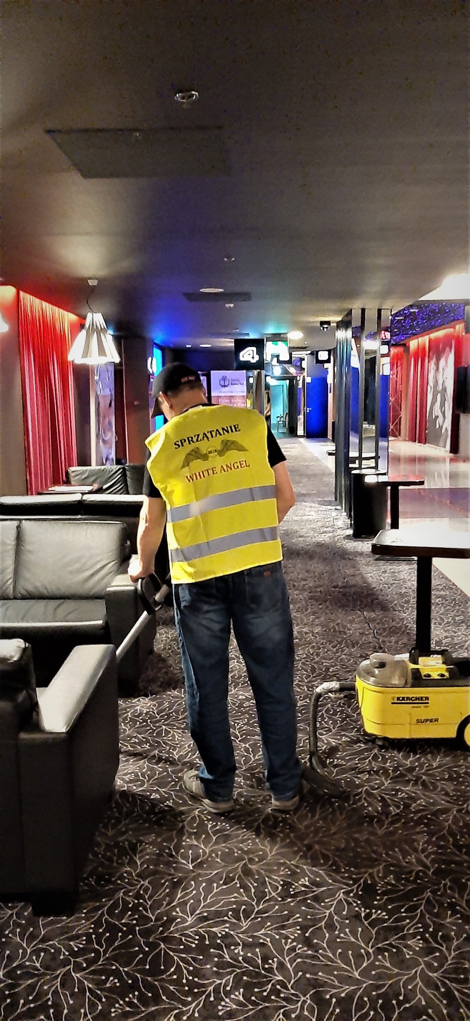 Osoba w żółtej kamizelce z napisem 'Sprzątanie White Angel' odkurza dywan w hotelowym lobby za pomocą odkurzacza Karcher Super.