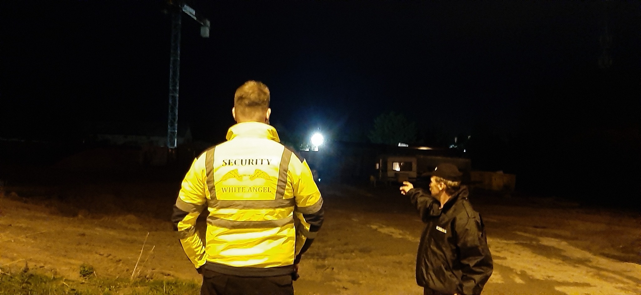 Dwóch ochroniarzy w ciemności, jeden w żółtej kurtce z napisem SECURITY i WHITE ANGEL na plecach, drugi w czarnej kurtce wskazuje palcem w oddali, w tle plac budowy z dźwigiem i oświetlony budynek.