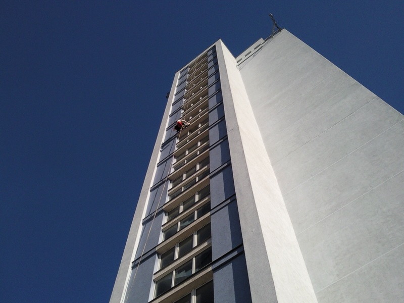 Pracownik na linie alpinistycznej, w trakcie pracy na wysokości, myjący okna wysokiego budynku z niebieskimi balkonami na tle błękitnego nieba.