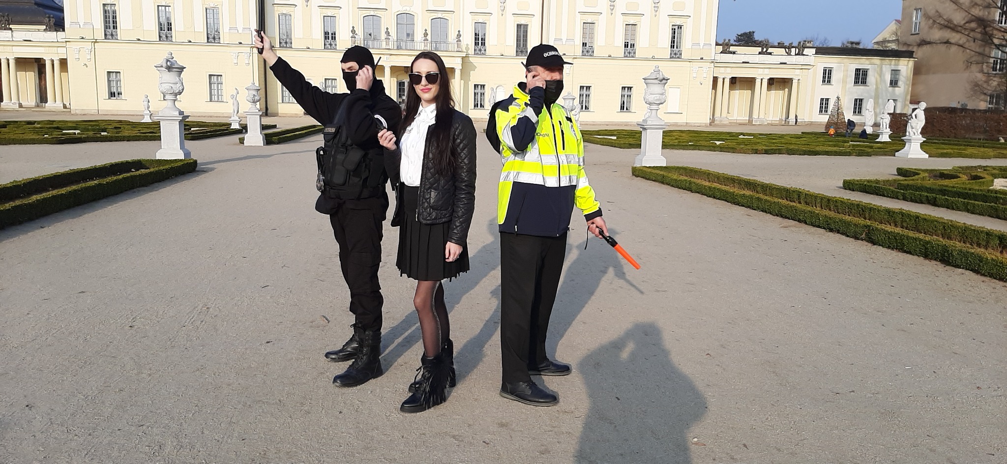 Trzech ochroniarzy w różnym umundurowaniu na tle pałacu i ogrodu, jeden z radiotelefonem, drugi z pałką.