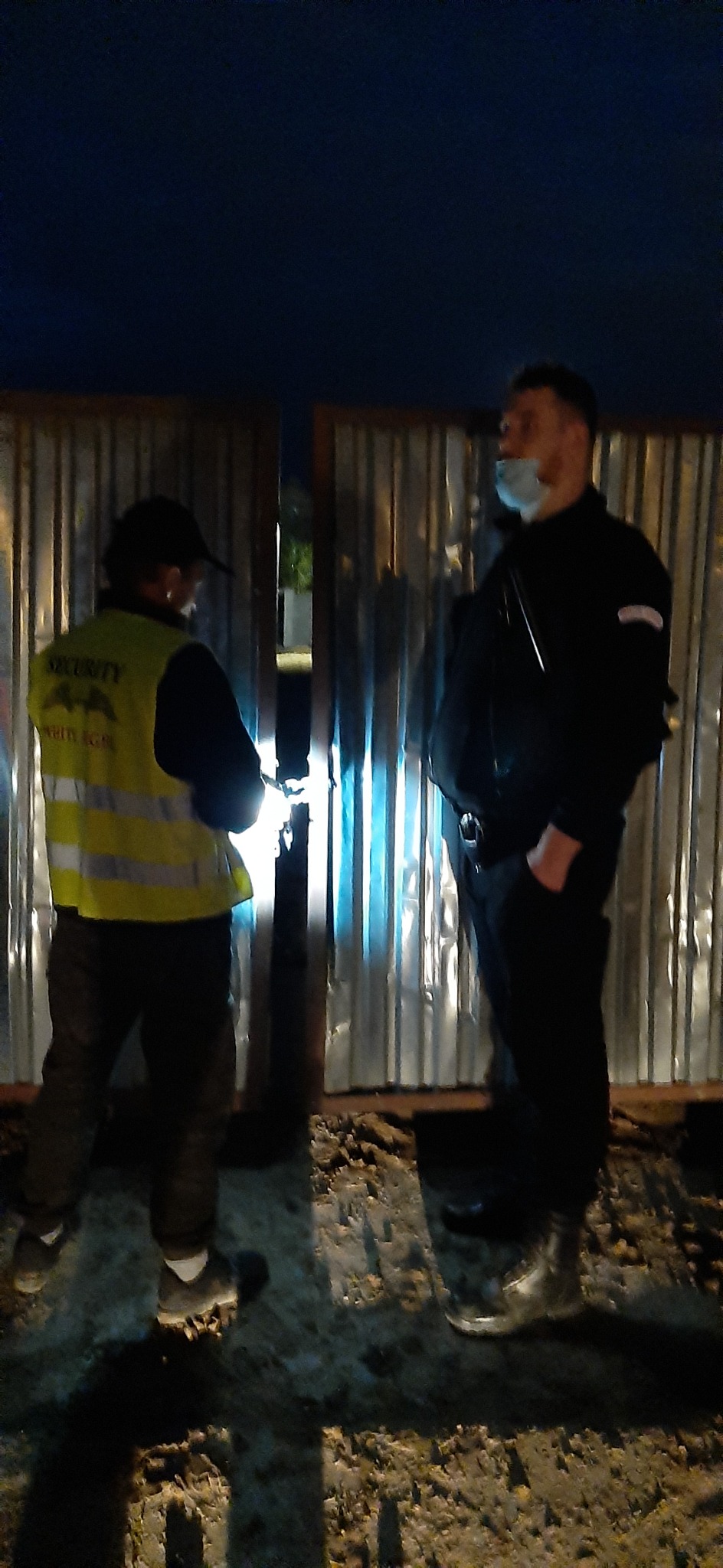 Dwóch mężczyzn w mundurach, jeden w kamizelce odblaskowej z napisem SECURITY, stoi przy metalowej bramie w słabym oświetleniu nocnym.