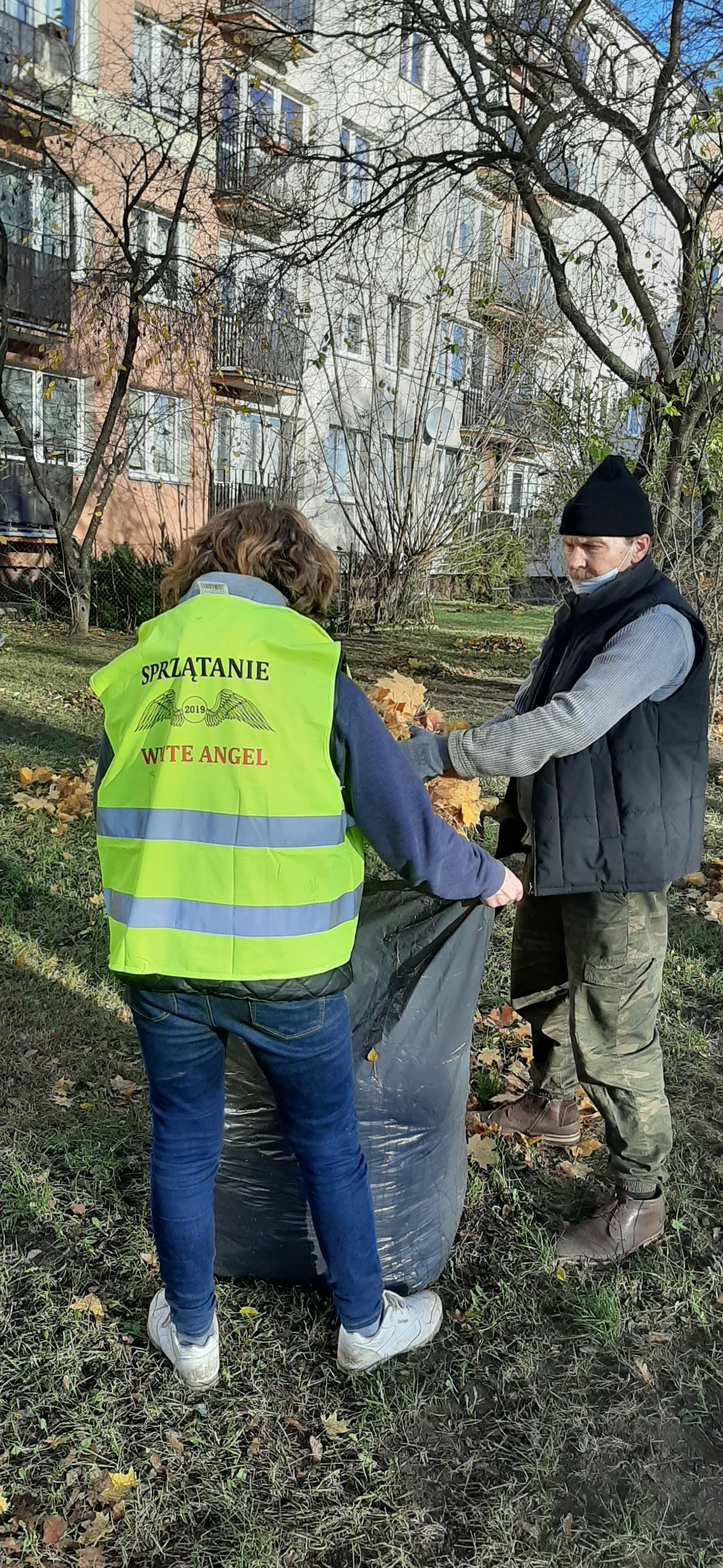 Dwie osoby zbierają opadłe liście do czarnego worka na tle bloku mieszkalnego. Jedna osoba ma na sobie żółtą kamizelkę z napisem 'Sprzątanie' i logo firmy 'Wite Angel'.