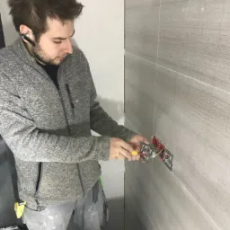 Elektryk w szarej bluzie podłącza gniazdko elektryczne na ścianie wyłożonej szarymi płytkami imitującymi drewno, używając śrubokręta z żółtą rękojeścią.