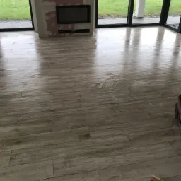 Świeżo ułożona, błyszcząca podłoga z płytek imitujących drewno w przestronnym salonie z dużymi oknami i przygotowanym miejscem na kominek.