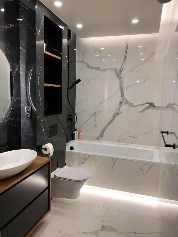 Nowoczesna łazienka z marmurowymi płytkami imitującymi żyły, podświetlaną wanną, umywalką nablatową na drewnianej szafce z czarnymi frontami i toaletą wiszącą.
