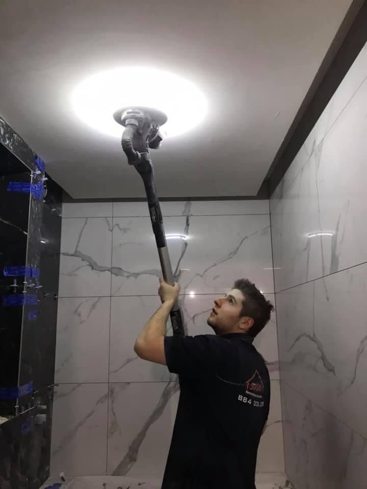 Mężczyzna szlifuje sufit w łazience wykończonej marmurowymi płytkami, używając szlifierki z długim uchwytem.