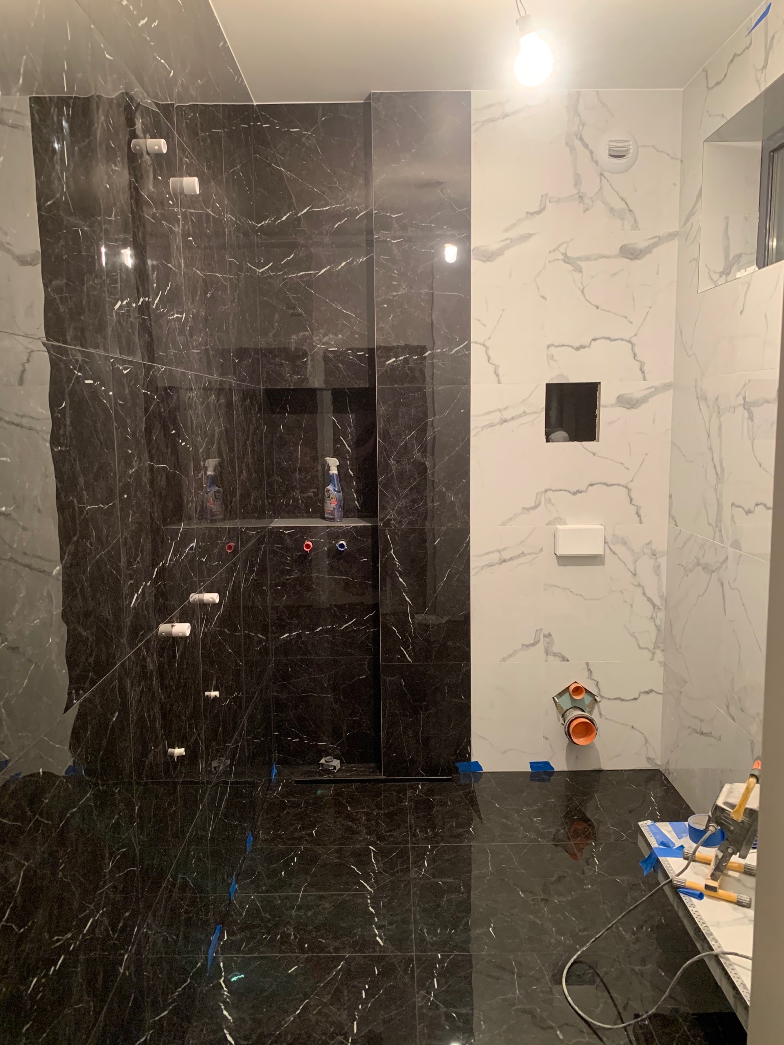 Łazienka w trakcie wykańczania, z czarnymi i białymi marmurkowymi płytkami, widoczne instalacje hydrauliczne, narzędzia glazurnicze i butelki ze środkami czyszczącymi na półce.