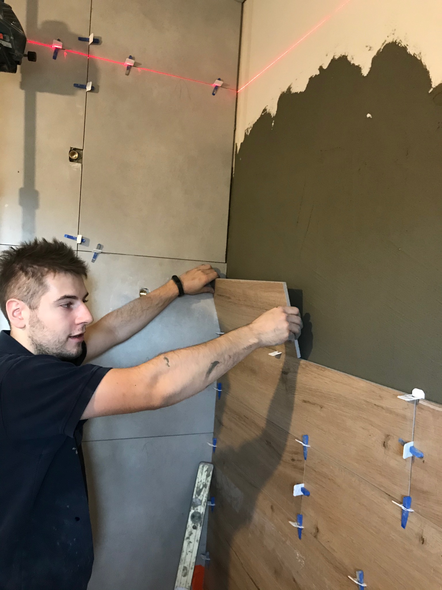 Glazurnik układa drewnopodobne płytki, używając poziomicy laserowej i klipsów do poziomowania, na ścianie z częściowo nałożonym klejem.