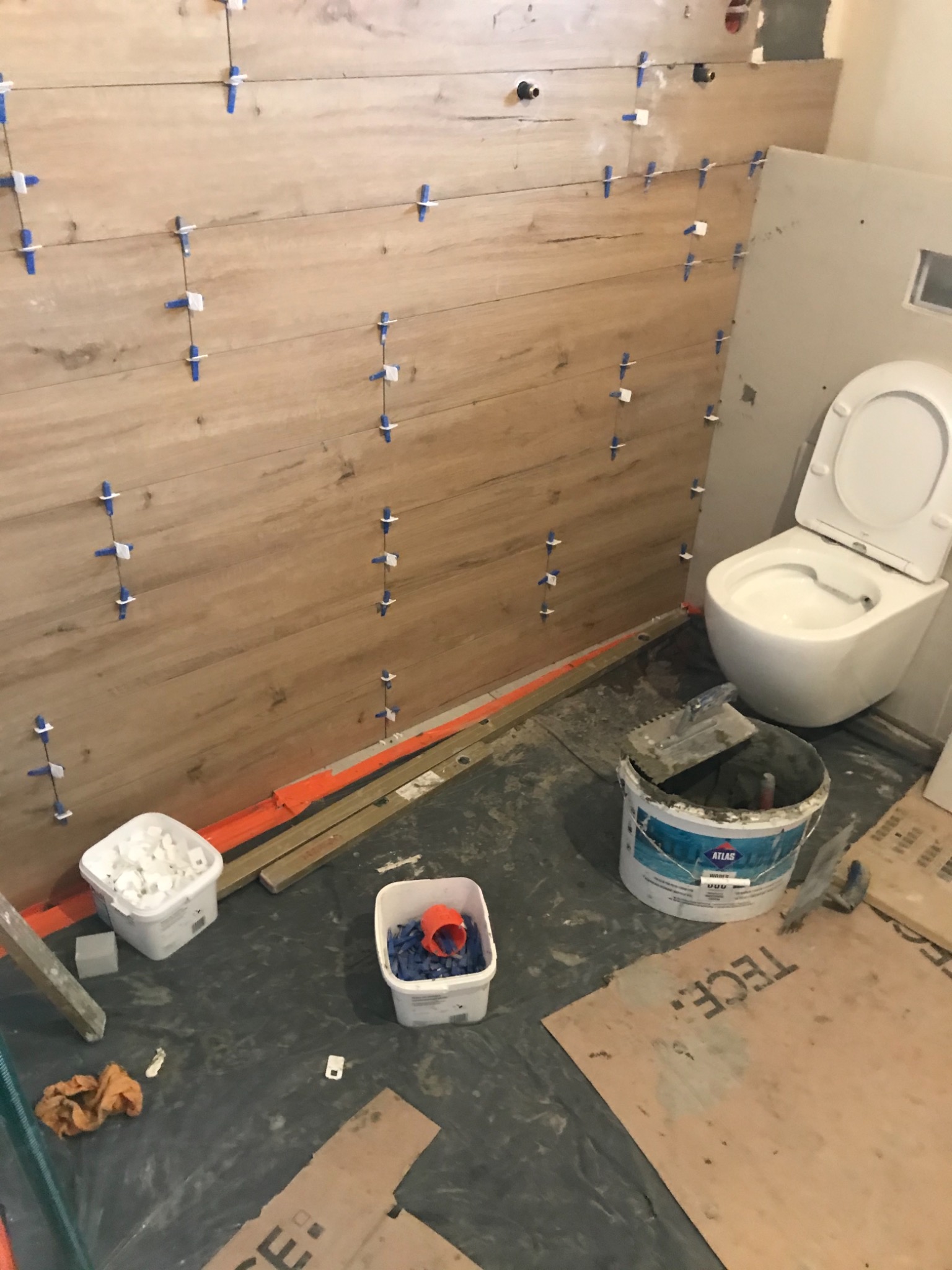 Ściana łazienki w trakcie układania płytek imitujących drewno, z użyciem niebieskich krzyżyków dystansowych, widoczna wisząca toaleta, wiadro z zaprawą klejową Atlas oraz pojemniki z krzyżykami...