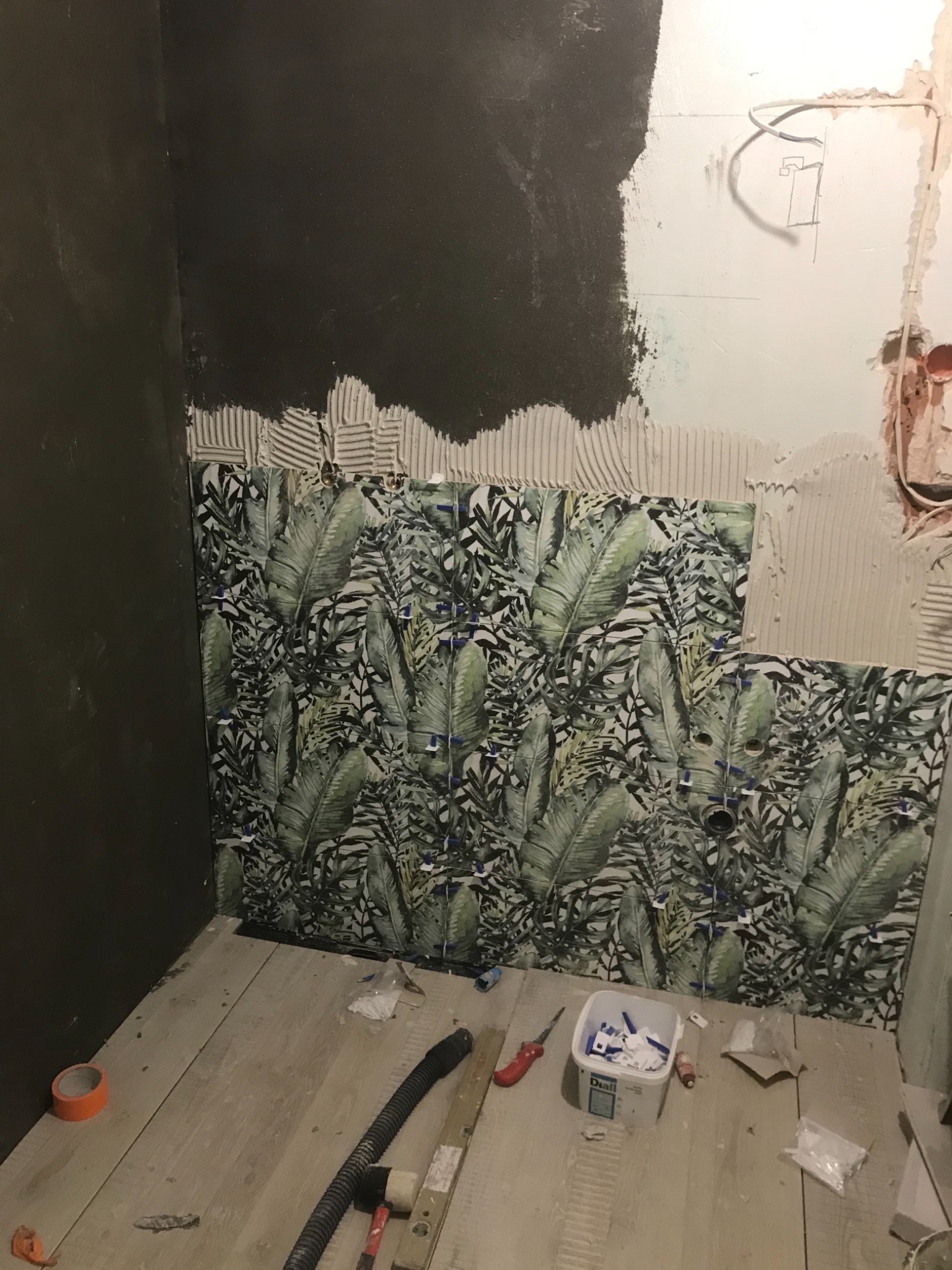 Ściana łazienki w trakcie układania płytek z motywem liści, widoczne narzędzia glazurnicze, klej oraz krzyżyki dystansowe.