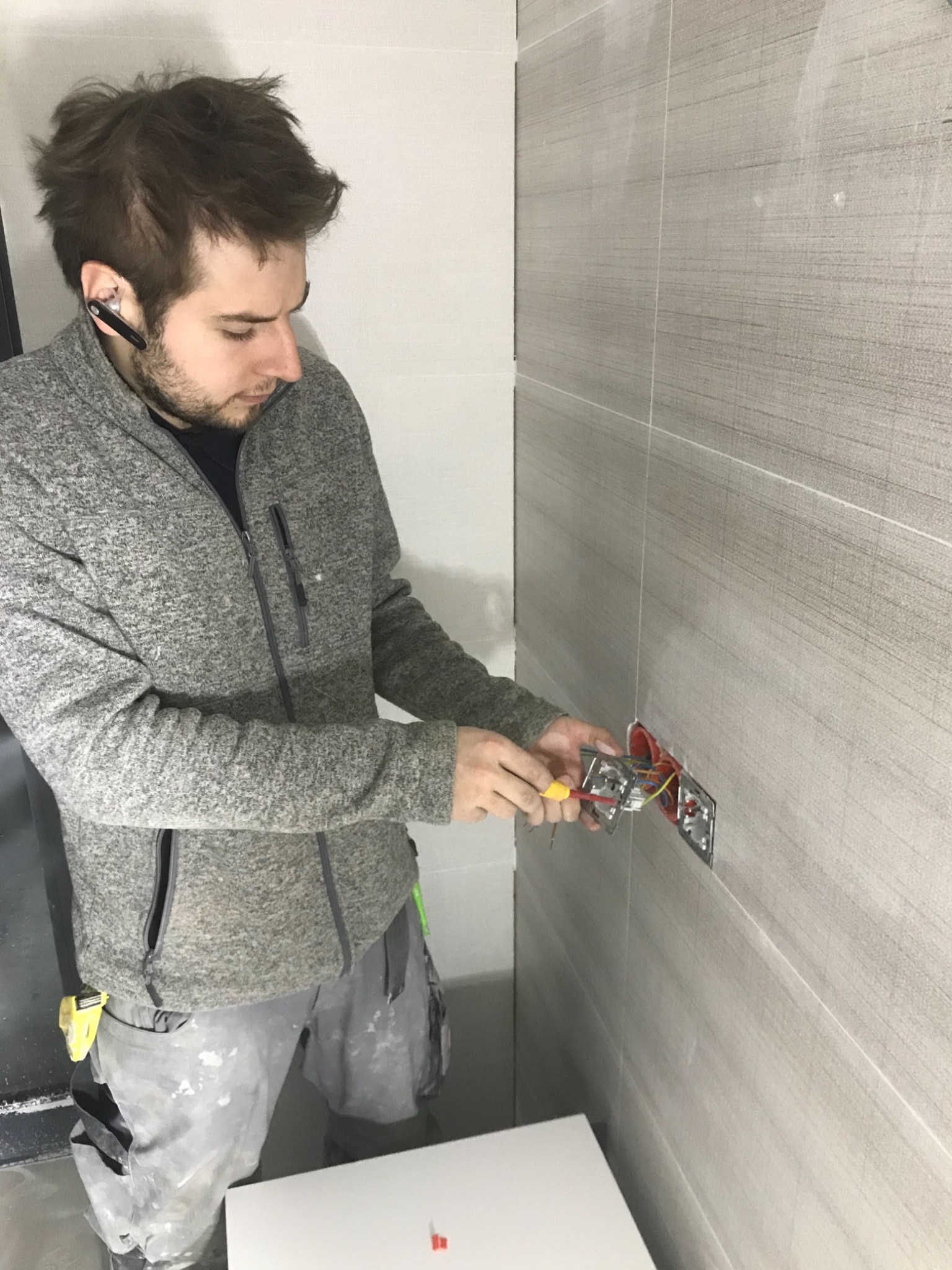 Elektryk w szarej bluzie podłącza gniazdko elektryczne na ścianie wyłożonej szarymi płytkami imitującymi drewno, używając śrubokręta z żółtą rękojeścią.