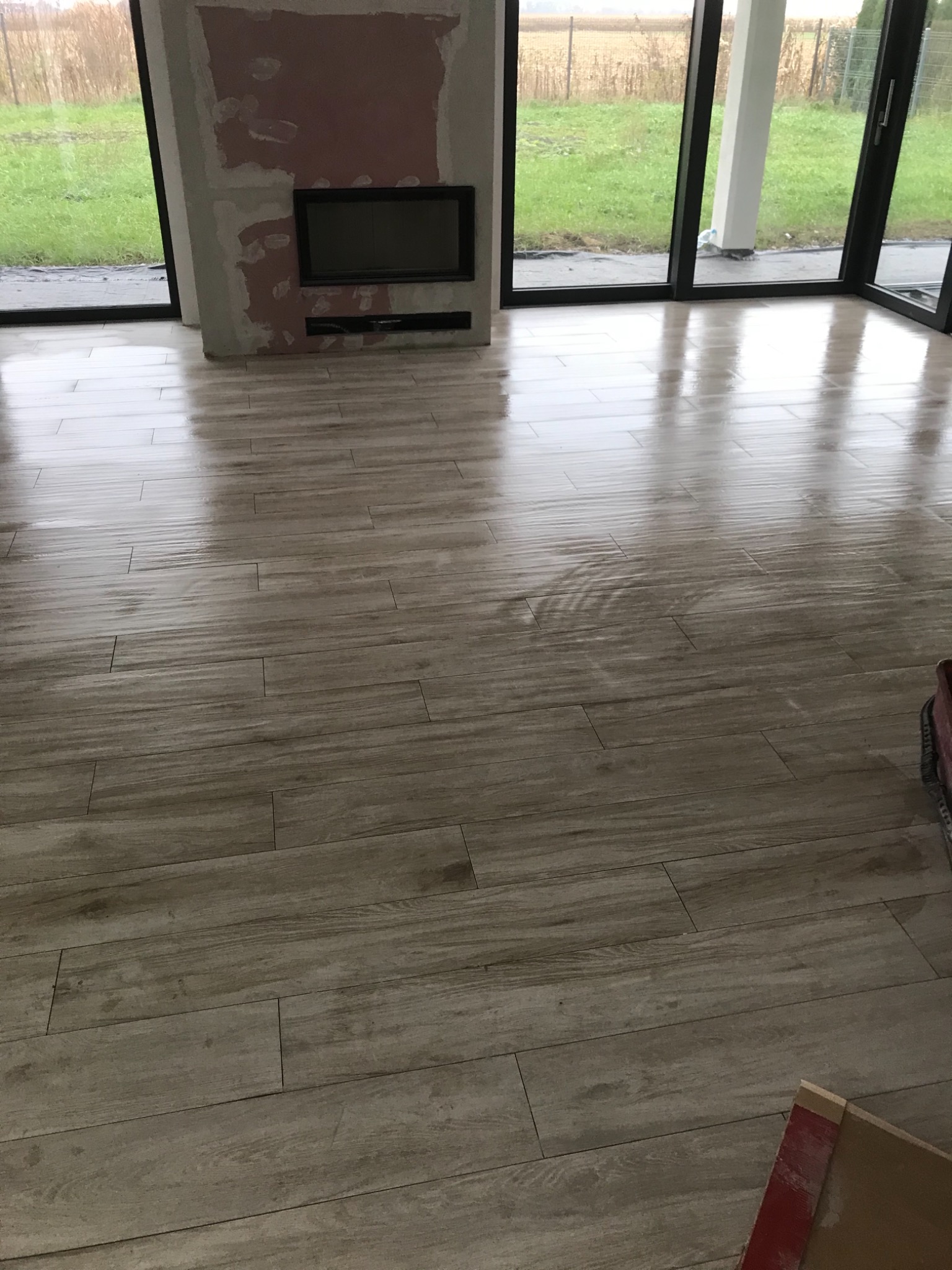 Świeżo ułożona, błyszcząca podłoga z płytek imitujących drewno w przestronnym salonie z dużymi oknami i przygotowanym miejscem na kominek.