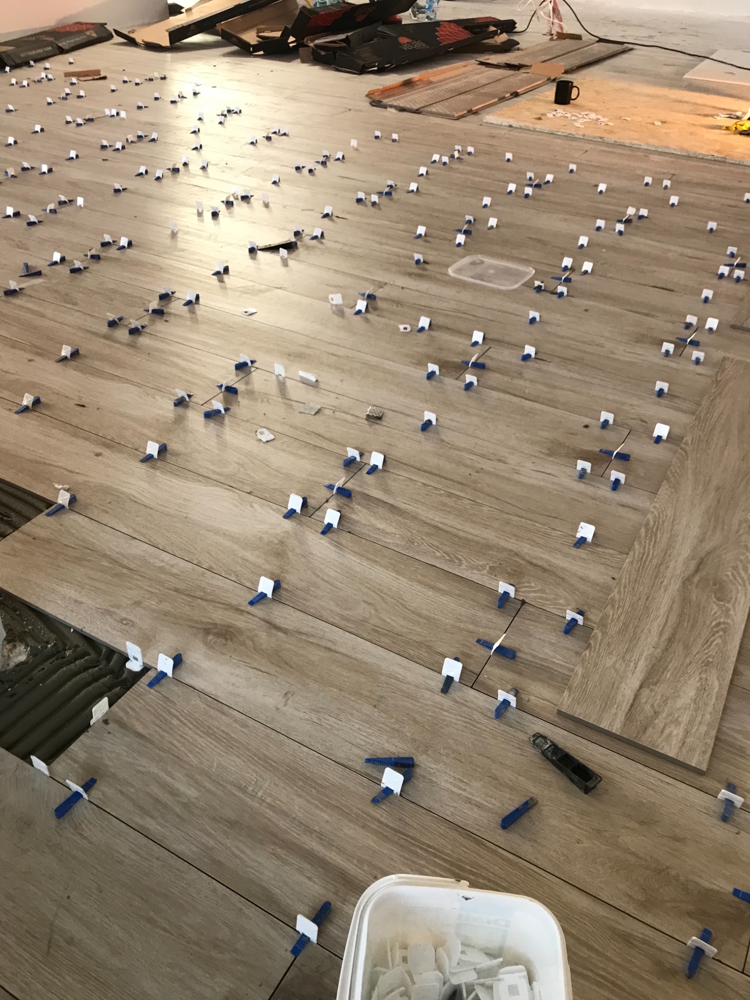 Układanie płytek podłogowych z użyciem systemu poziomowania płytek, widoczne liczne niebiesko-białe klipsy i kliny, wiadro z zapasem klipsów, oraz fragment ułożonej podłogi z widoczną fugą.