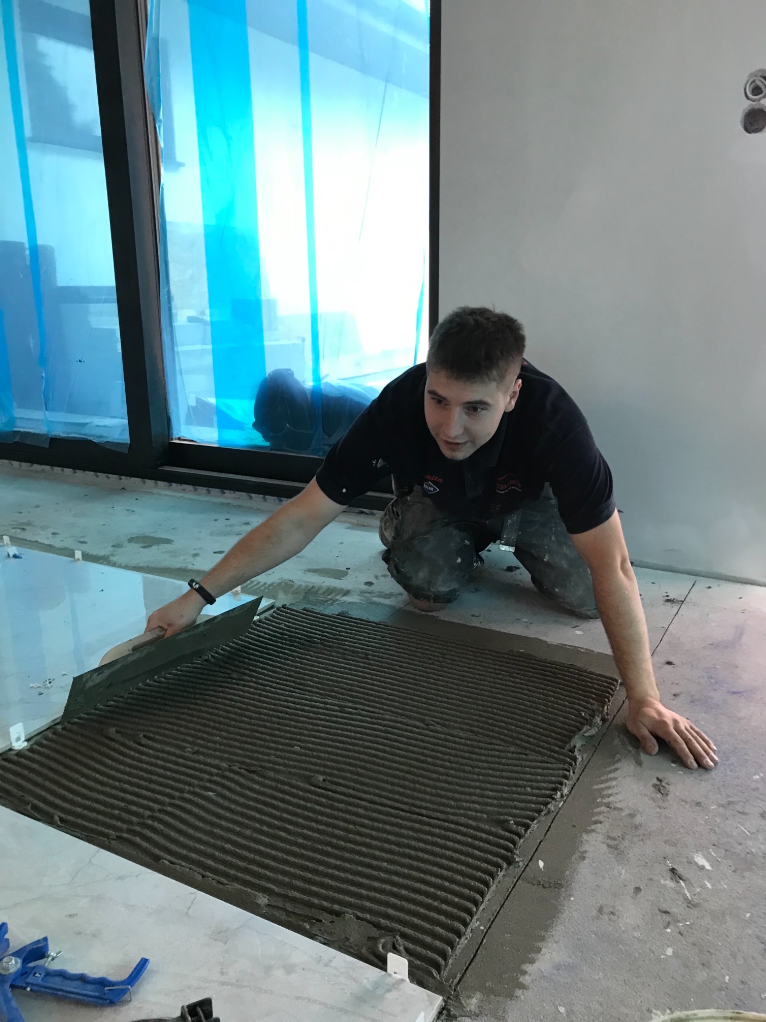 Mężczyzna w ciemnej koszulce, na kolanach, rozprowadza klej grzebieniem na podłodze przygotowanej pod płytki; w tle okno z niebieską folią ochronną.