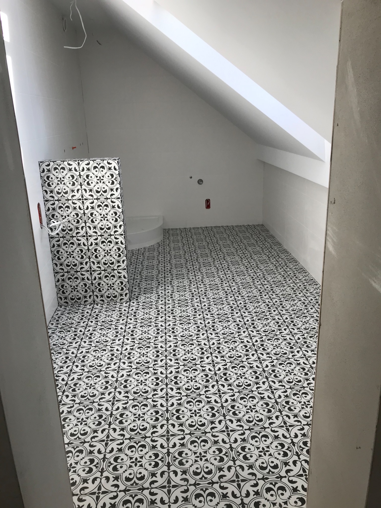 Łazienka na poddaszu w trakcie wykańczania, z biało-czarną wzorzystą podłogą i fragmentem kabiny prysznicowej, widoczne niedokończone ściany i wystające kable.