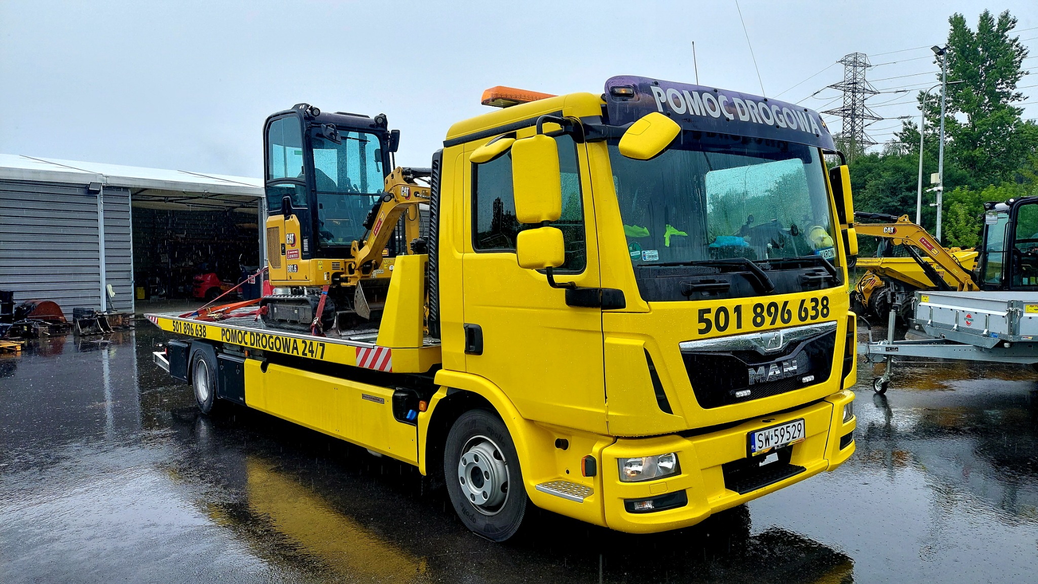 Żółty pojazd pomocy drogowej MAN z mini-koparką CAT na platformie, widoczne numery telefonu i napis 'POMOC DROGOWA 24/7', mokry asfalt odbija światło, w tle budynek magazynowy i słupy wysokiego...