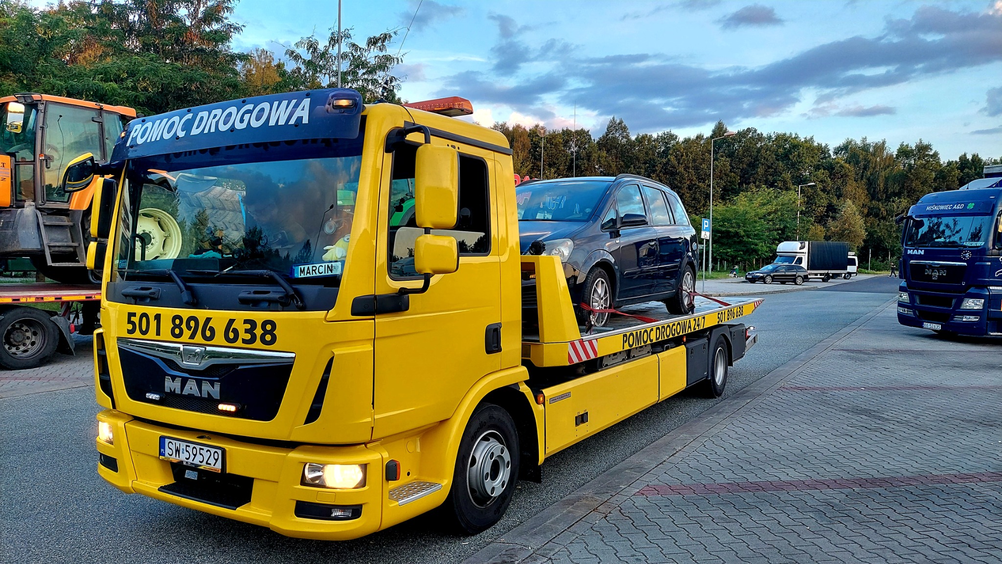 Żółta laweta MAN z czarnym samochodem osobowym na platformie, zaparkowana na parkingu z kostki brukowej, w tle inne pojazdy i drzewa pod wieczornym niebem.