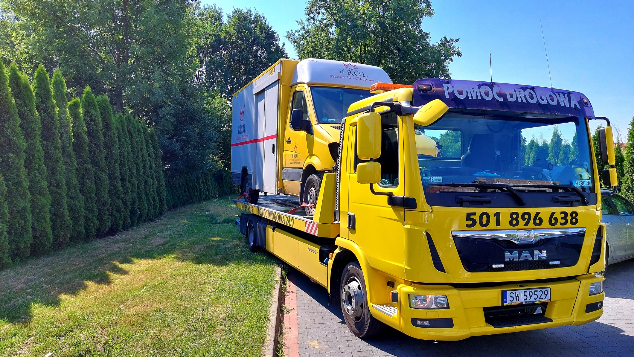 Żółty pojazd pomocy drogowej MAN z napisem 'POMOC DROGOWA' na osłonie przeciwsłonecznej, przewożący na platformie biało-żółty samochód dostawczy z logo piekarni 'KROL'.