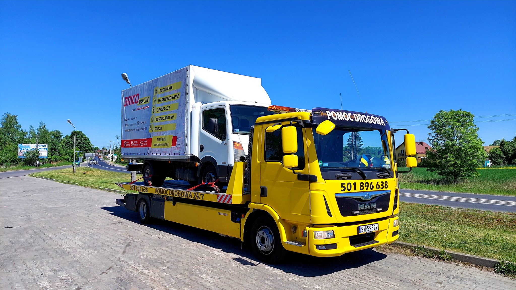 Żółta ciężarówka pomocy drogowej z napisem 'POMOC DROGOWA' przewozi na platformie biały samochód dostawczy z reklamą 'Brico Marche' na tle błękitnego nieba i zielonej okolicy.