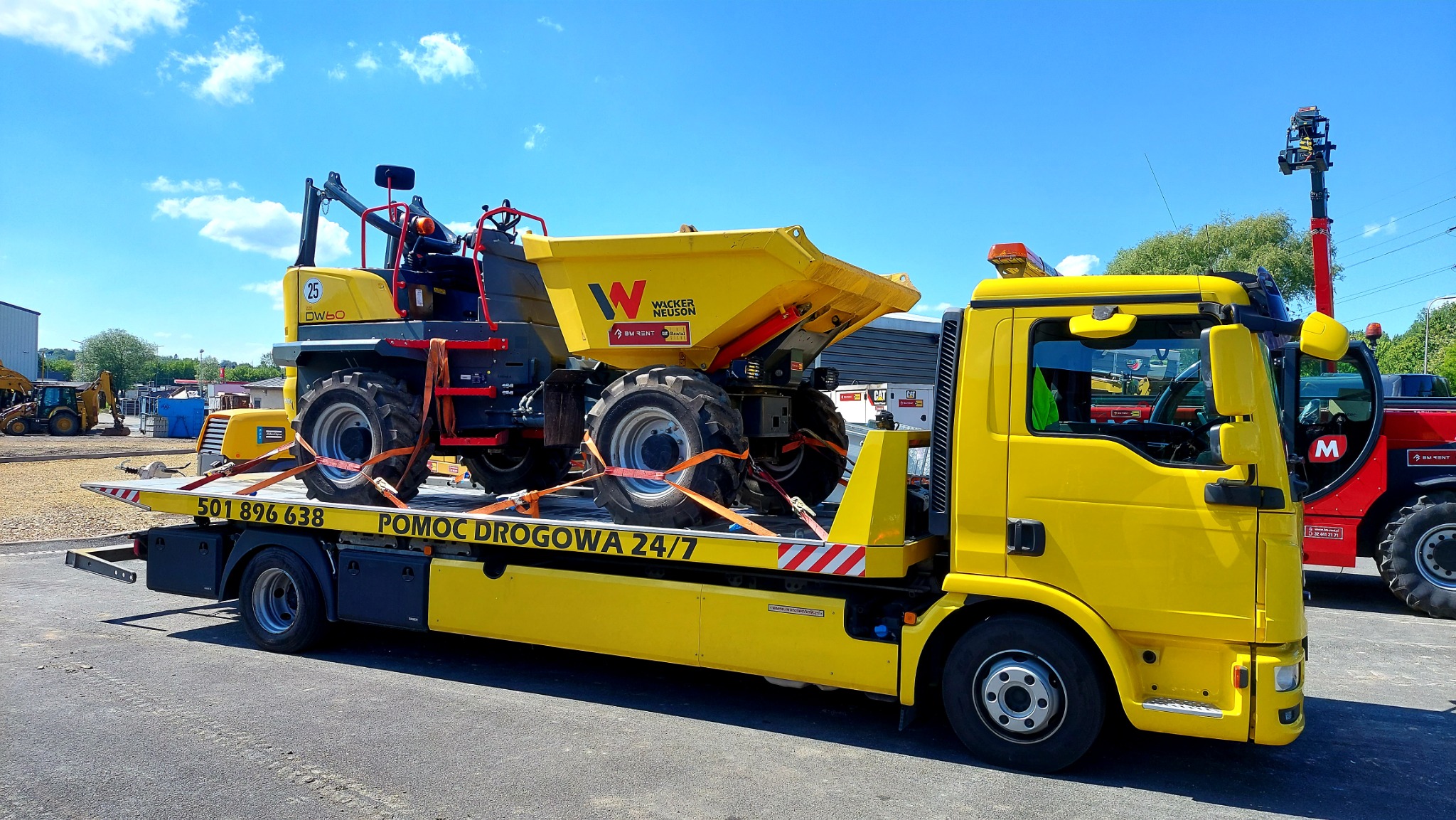 Żółty samochód pomocy drogowej z platformą transportową przewozi żółtą wywrotkę budowlaną Wacker Neuson, zabezpieczoną pomarańczowymi pasami. Widoczne numery kontaktowe i napis 'Pomoc Drogowa 24/7'.