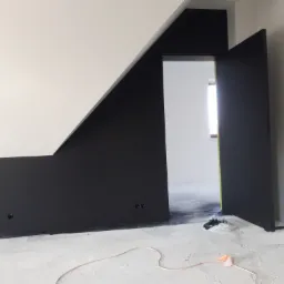 Ściana ze skosem pomalowana na czarno i biało, otwarte czarne drzwi, widoczne ślady farby na podłodze, zabezpieczone gniazdka elektryczne.