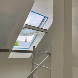 Wnętrze w Siedliszczach w trakcie malowania, widoczny fragment rusztowania i okno dachowe zabezpieczone taśmą malarską, ściany w trakcie prac malarskich.