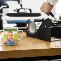 Biały kubek z nadrukowanym zdjęciem uśmiechniętego dziecka, umieszczony obok prasy termicznej do kubków obsługiwanej przez osobę w koszuli z podwiniętymi rękawami, na drewnianym stole.