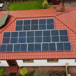 Widok z góry na dom z czerwoną dachówką, na której zamontowano panele słoneczne; dodatkowo widoczny kolektor słoneczny obok komina.