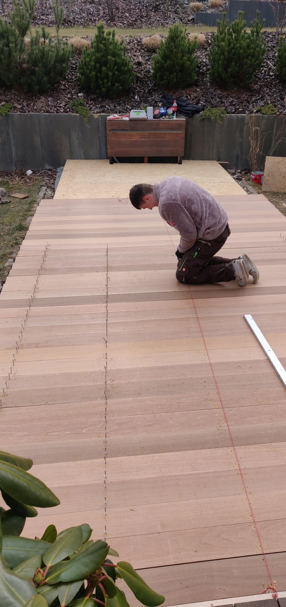 Mężczyzna klęczy na drewnianym tarasie, wyznaczając linię za pomocą sznurka i gwoździ, w tle widoczne krzewy i drewniana skrzynia.