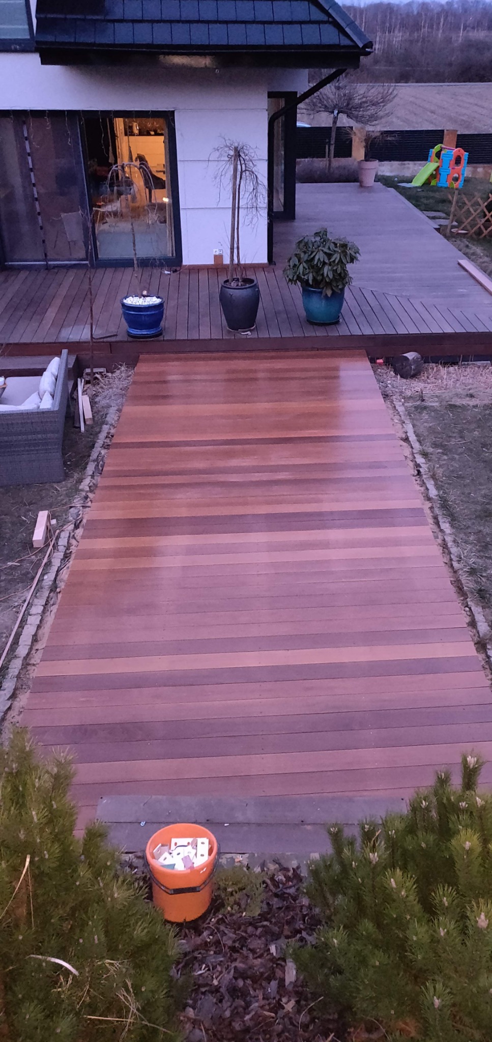 Świeżo wykończony drewniany taras z desek kompozytowych prowadzący do domu, z doniczkami i elementami placu zabaw w tle.