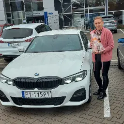 Kobieta z prezentem stoi obok białego BMW na tle salonu samochodowego Bemo Motors.