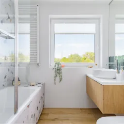 Jasna łazienka z wanną obudowaną płytkami lastryko, drewnianą szafką pod umywalką i dużym oknem z widokiem na zieleń.