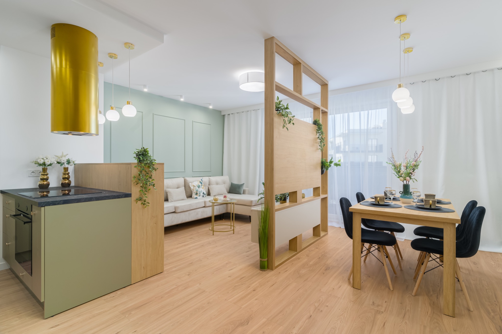 Jasne, przestronne wnętrze z drewnianą podłogą, zielonymi akcentami i złotymi detalami, obejmujące aneks kuchenny z nietypowym, złotym okapem cylindrycznym, stół jadalny z czarnymi krzesłami...