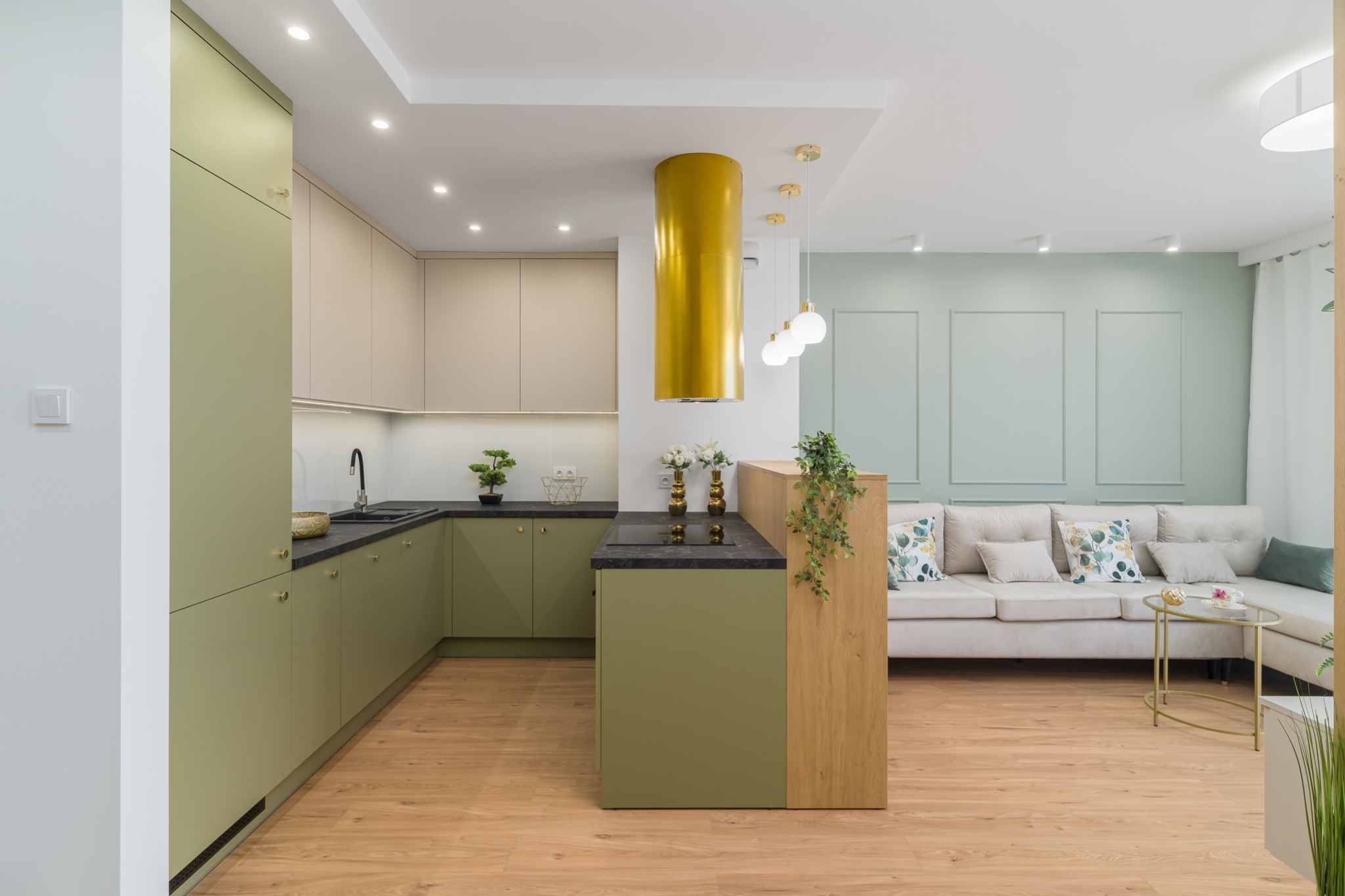 Nowoczesna kuchnia otwarta na salon, z zielonymi szafkami, czarnym blatem, złotym okapem cylindrycznym i drewnianą podłogą.