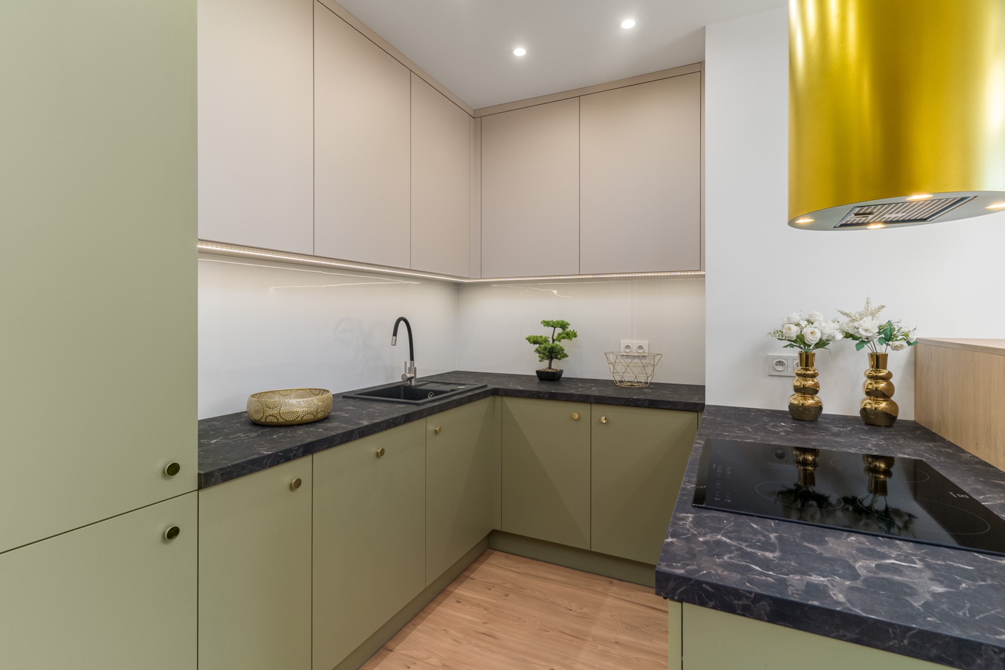 Nowoczesna kuchnia w odcieniach zieleni i beżu z czarnym blatem, złotym okapem i drewnianą podłogą, oświetlona ledowym światłem podszafkowym i punktowymi lampami sufitowymi.