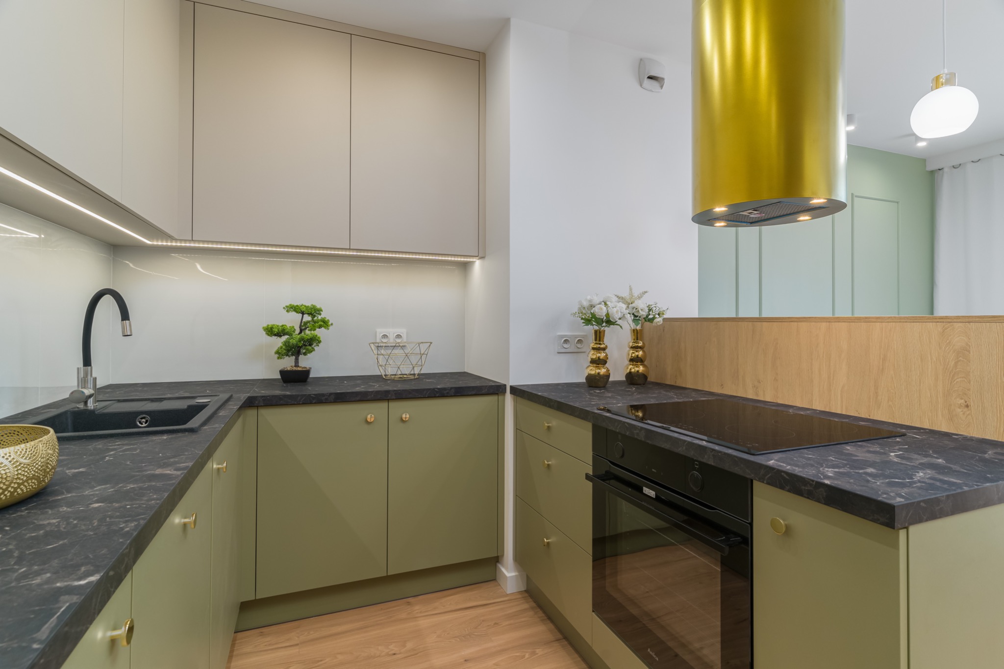 Nowoczesna kuchnia w odcieniach zieleni i beżu z czarnym blatem, złotymi akcentami i cylindrycznym, złotym okapem.
