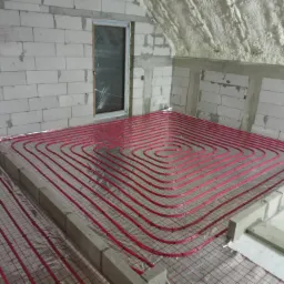 Poddasze w trakcie wykończenia, widoczne ogrzewanie podłogowe w postaci czerwonych rurek ułożonych spiralnie na folii izolacyjnej, ściany z białych bloczków betonowych i fragment ocieplonego pianą...