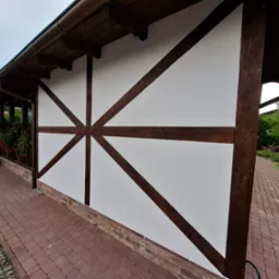 Biała elewacja budynku z drewnianą konstrukcją szkieletową w brązowym kolorze, widok z boku, z fragmentem chodnika z kostki brukowej i zieleni w tle.