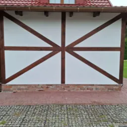 Biała ściana szachulcowa z brązowymi belkami, ceglana podmur&oacute;wka i czerwona kostka brukowa. W tle trawnik i plac zabaw. Detal architektury.