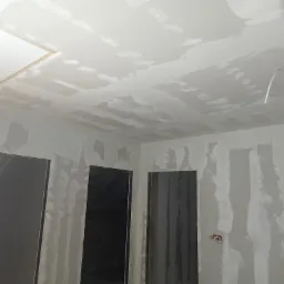 Wnętrze w trakcie wykańczania: surowe ściany i sufit pokryte szpachlą, widoczne otwory drzwiowe bez ościeżnic, wystające przewody elektryczne, klapa rewizyjna w suficie.