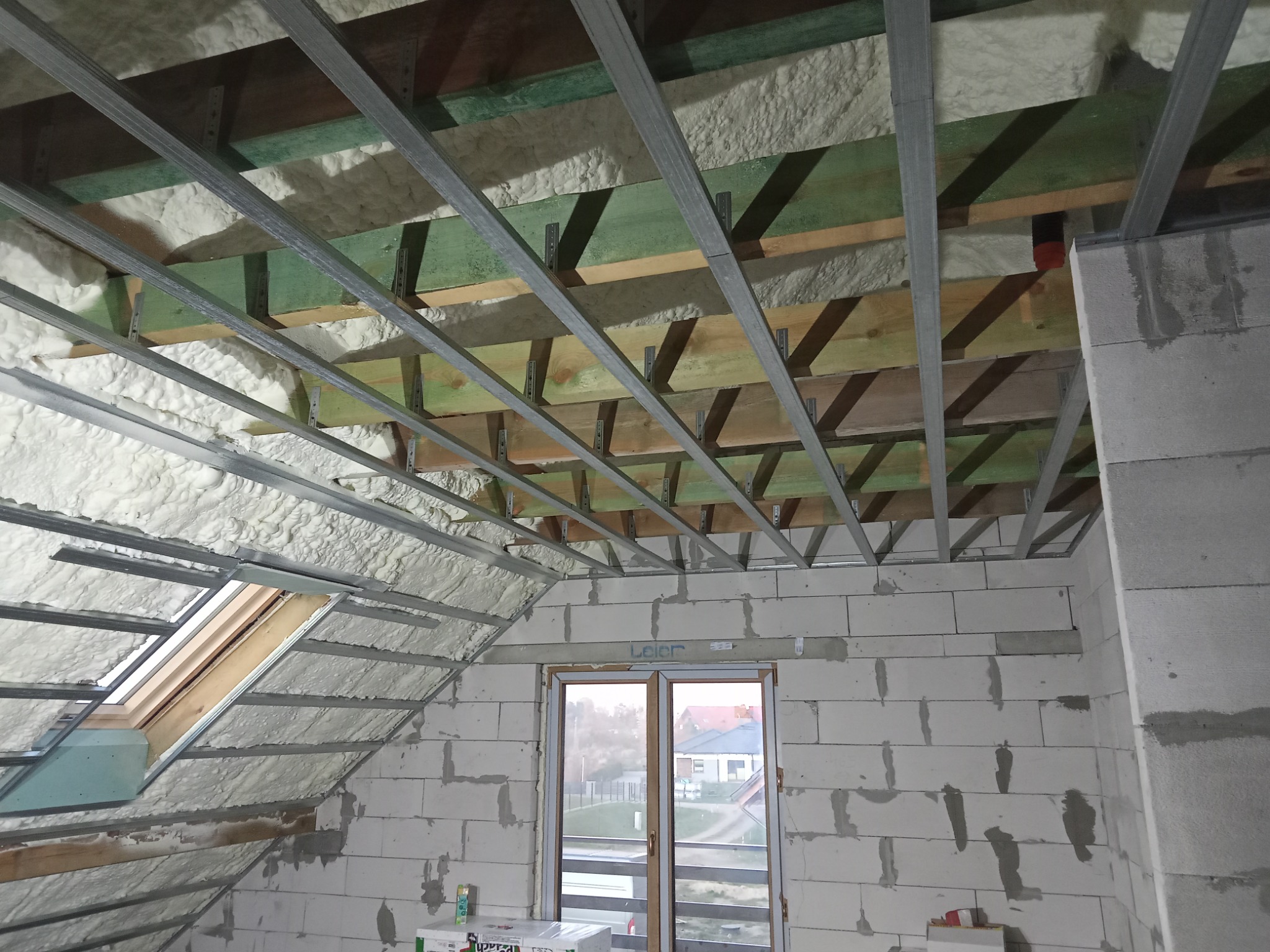 Poddasze w trakcie ocieplania pianą, widoczne drewniane belki, metalowa konstrukcja sufitu podwieszanego oraz okno dachowe i okno w ścianie szczytowej.