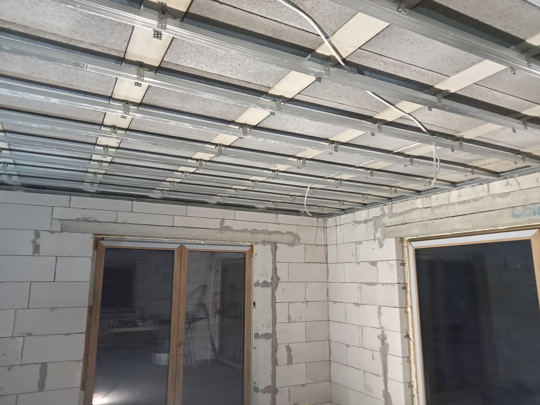 Stelaż metalowy podwieszanego sufitu z płytami w surowym stanie, widoczne przewody, ściany z białych bloczków.