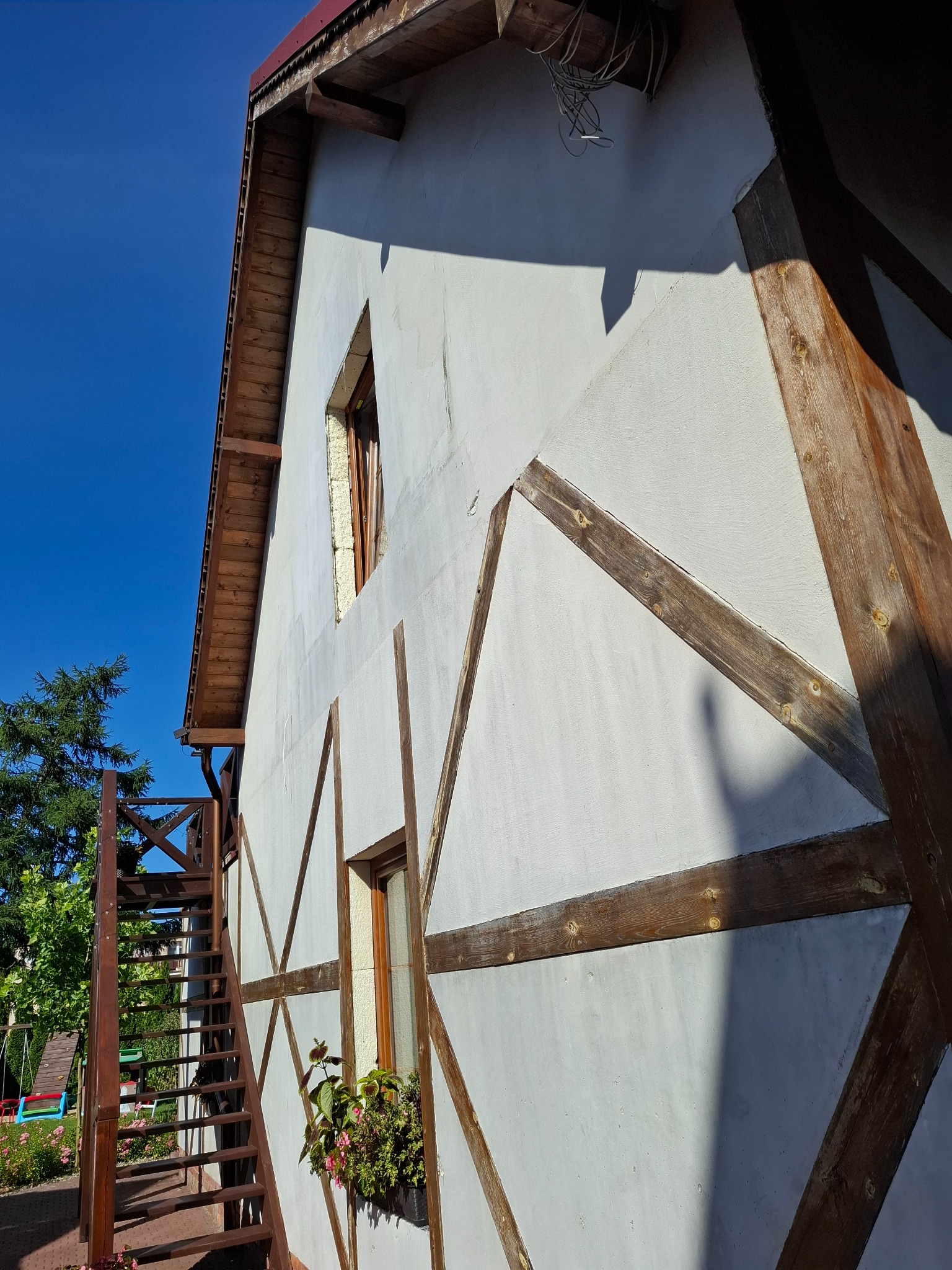 Boczny widok domu z białą elewacją i drewnianymi elementami konstrukcyjnymi. Widoczne okna, schody zewnętrzne i fragment ogrodu. Słoneczny dzień.