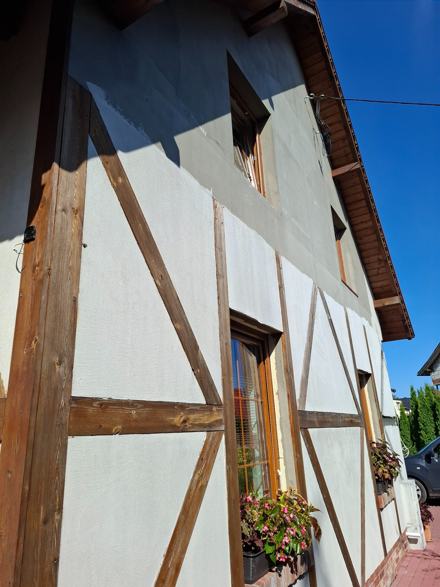 Elewacja domu w stylu szachulcowym po renowacji, z drewnianymi belkami i białymi polami. Widoczne okna z kwiatami i fragment dachu. Słoneczny dzień.