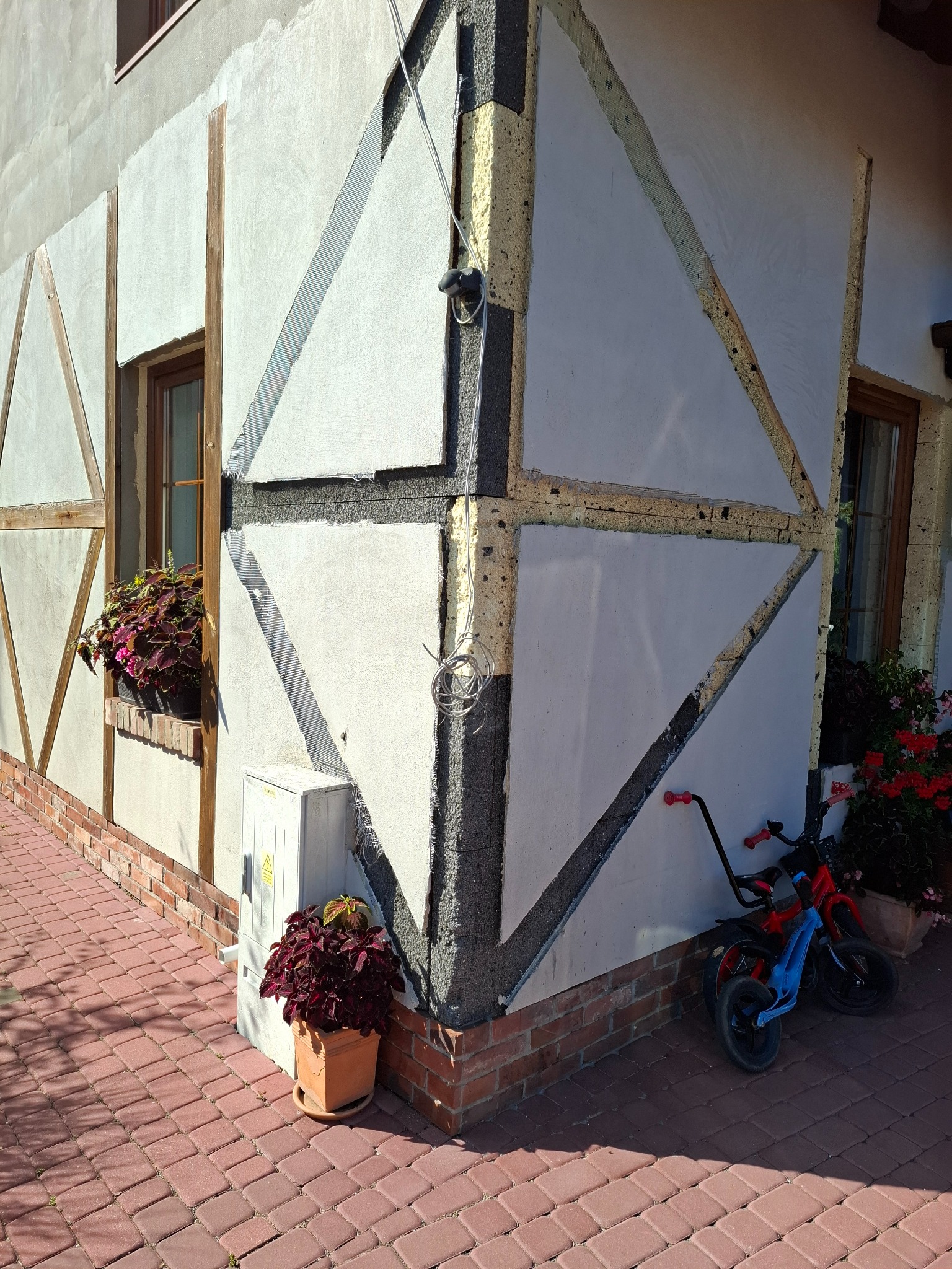 Fragment elewacji domu w trakcie ocieplania, widoczne elementy konstrukcyjne i siatka zbrojeniowa. Obok stoi rower dziecięcy i doniczka z kwiatami.